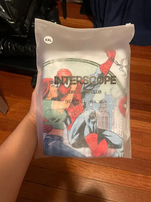 A custom poly mailer held in someone’s hand displays stylized superhero-themed artwork and the text 'INTERSCOPE DREAMERS CLUB' with additional unreadable script.